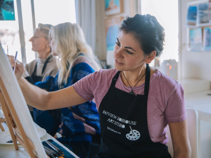 A woman smiles as she paints on a canvas against a wooden table top easel at a group painting class